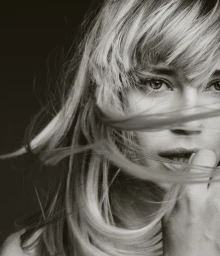Monochrome close-up portrait of a woman with fringe bangs and strands of hair blowing across her face against a dark background.