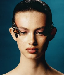 Close-up beauty portrait of a woman with slicked-back hair, pink eyeshadow, and monarch butterfly wings applied near her eyes.
