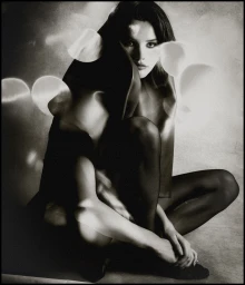 Monochrome portrait of a woman seated wearing black stockings and draped dark fabric with artistic circular lens flare effects.