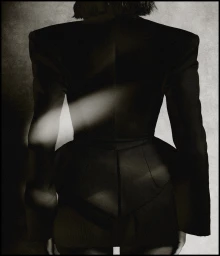 Back view of a person wearing a structured black blazer with exaggerated shoulders and cinched waist against a textured backdrop.