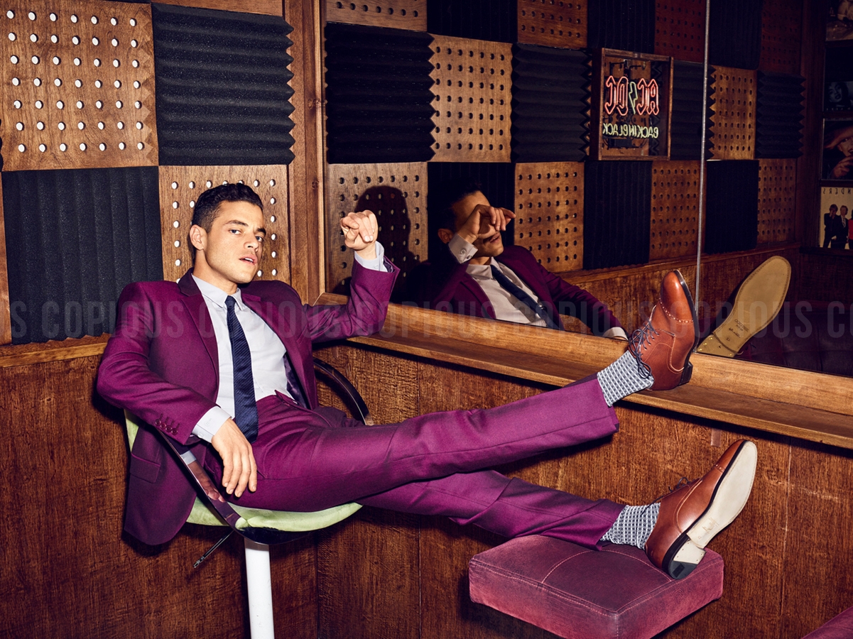 Rami Malek lounges in a purple suit in a wood-paneled music studio with soundproofing. Reflected image visible in the mirror.