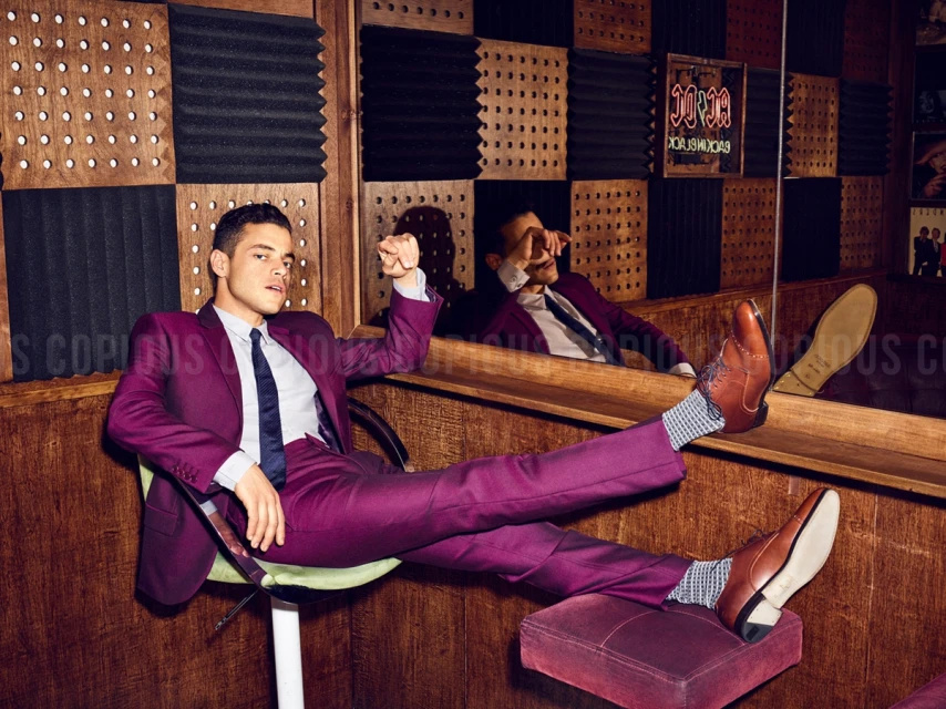 Rami Malek lounges in a purple suit in a wood-paneled music studio with soundproofing. Reflected image visible in the mirror.