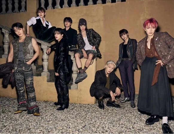 Group of eight young adult male fashion models posing together outdoors, wearing a variety of modern clothing, with a neutral architectural background.