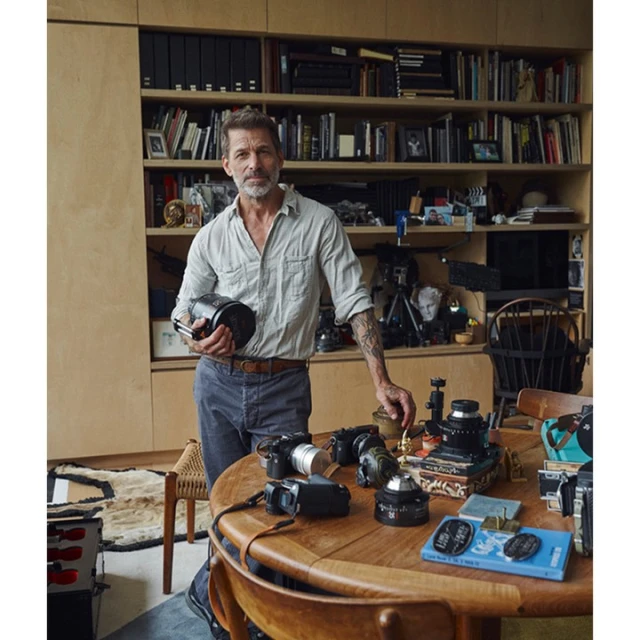Man with grey hair poses indoors, holding a camera lens. A table displays camera equipment, with shelves and books in the background.