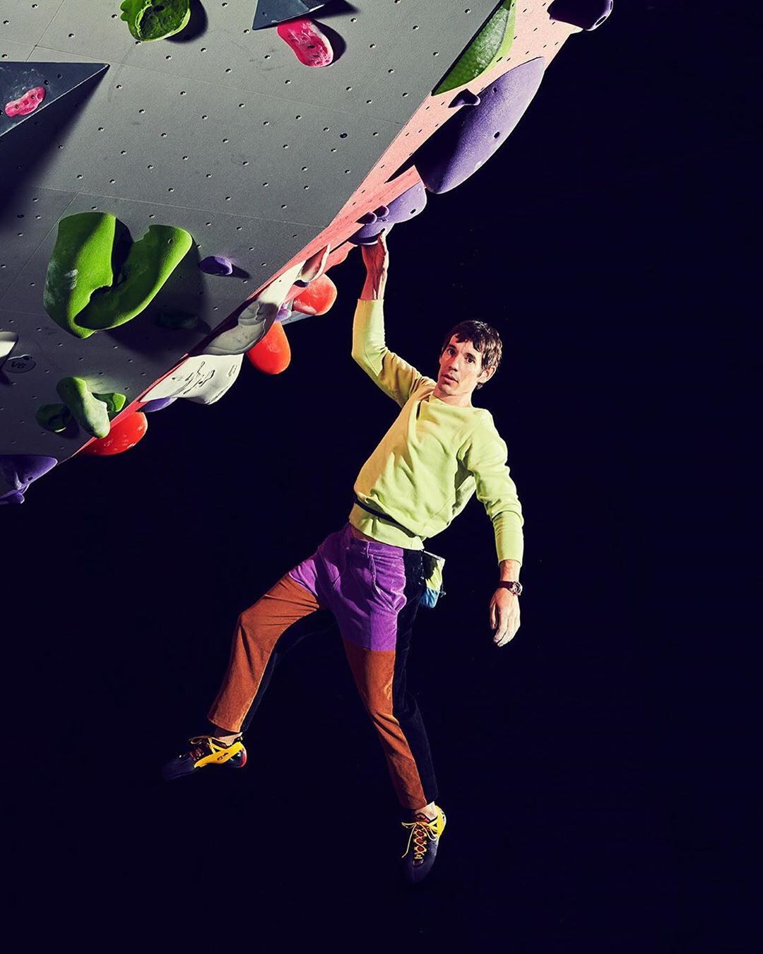 An adult rock climber on an indoor rock wall, wearing colourful athletic clothing and climbing shoes, against a dark background.