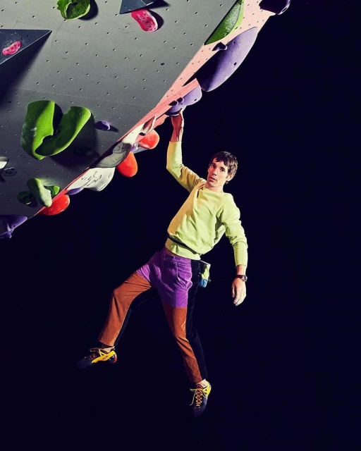An adult rock climber on an indoor rock wall, wearing colourful athletic clothing and climbing shoes, against a dark background.