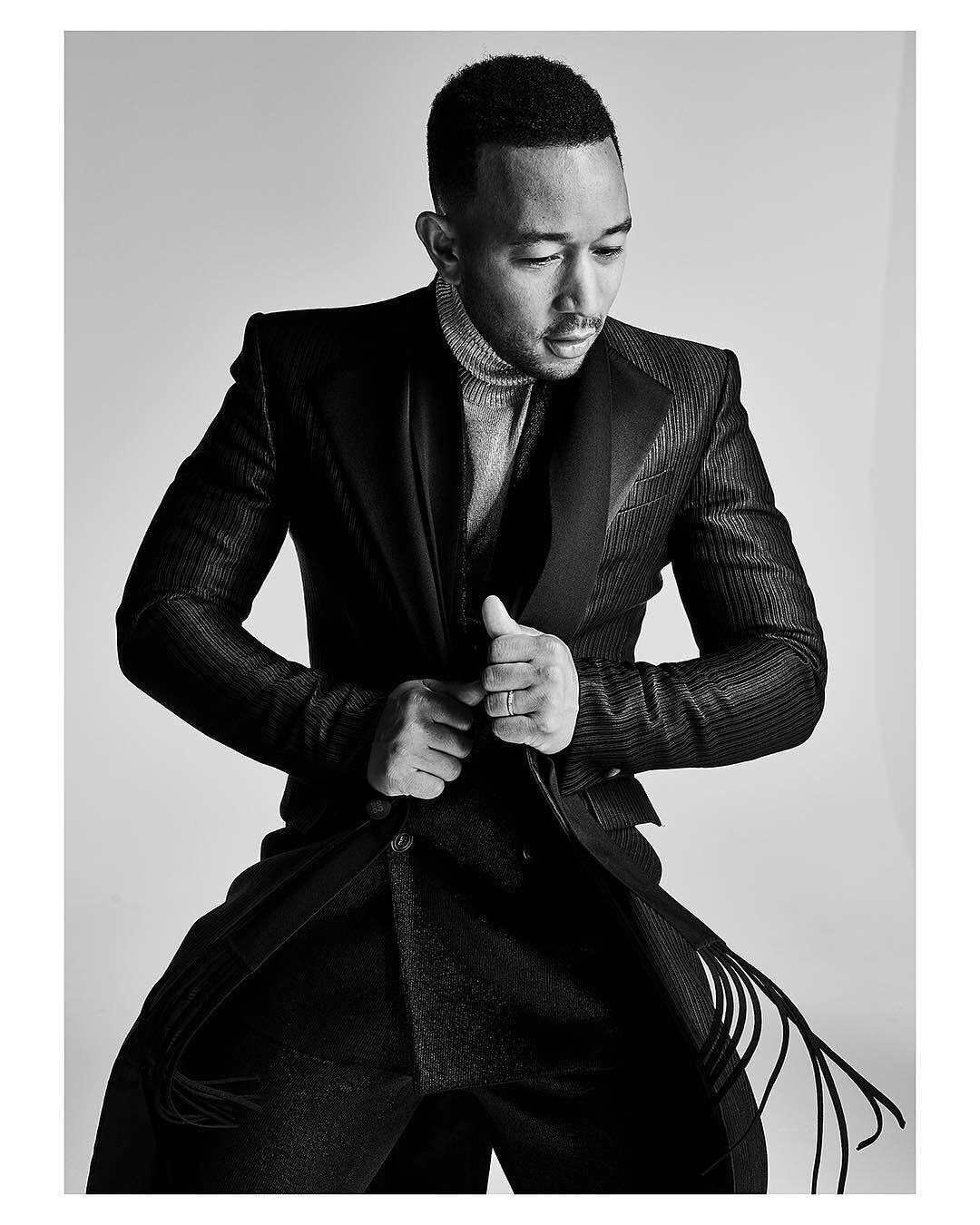 Black and white portrait of John Legend wearing a dark suit and light turtleneck, looking down with hands clasped in front of him.