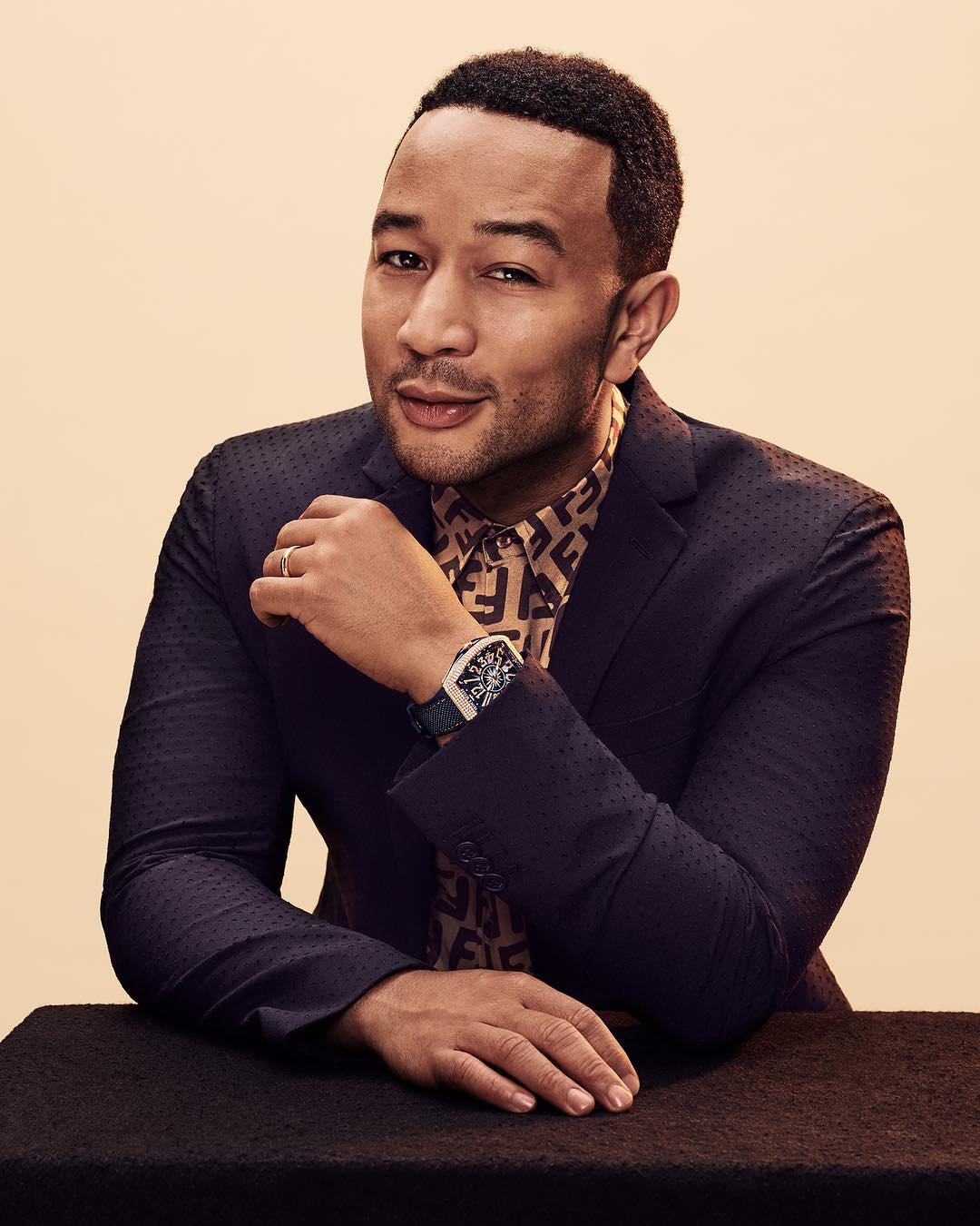 Portrait of singer John Legend in a patterned shirt and black blazer in a studio setting, looking towards the camera with a slight smile.