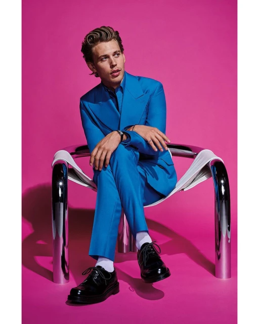 Portrait of stylish man, in a turquoise suit, sitting on a chrome chair against a pink backdrop, exuding confidence and modern elegance.