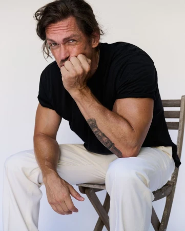 Man seated on a wooden folding chair wearing a black t-shirt and white trousers against a plain white background.