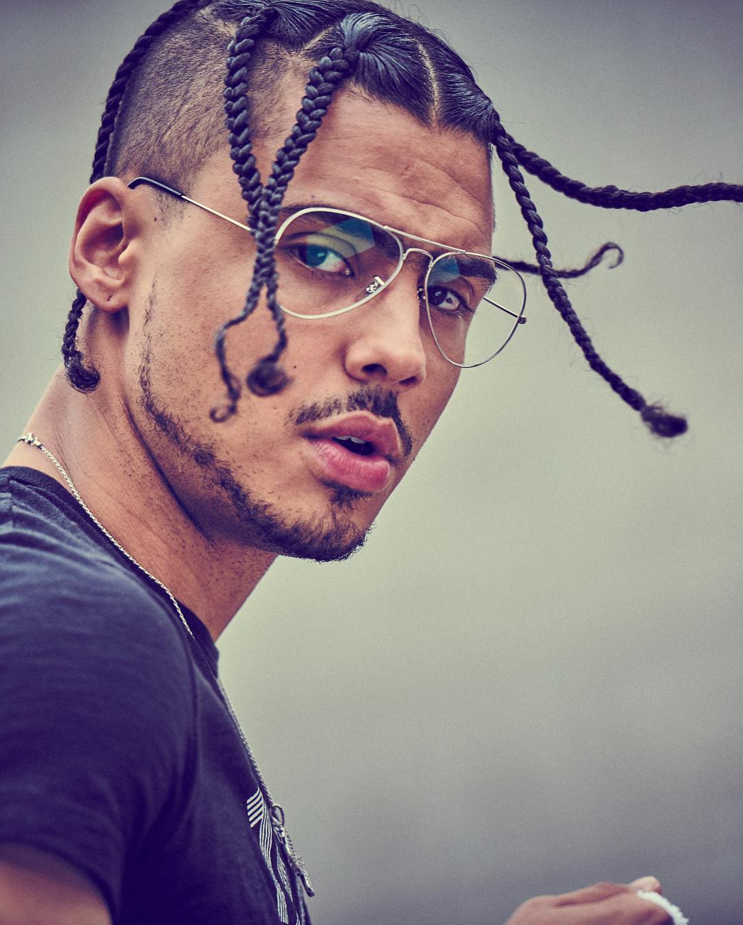 Portrait of a young adult male with braids and aviator glasses looking at the camera against a blurred gray background.