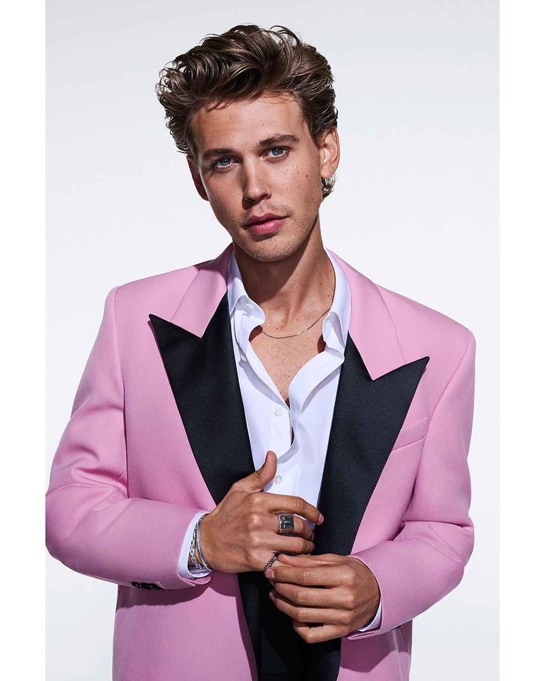 Portrait of a young adult male with wavy brown hair in a pink tuxedo jacket and white shirt, posing against a bright white backdrop.