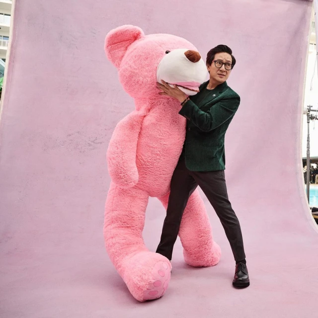 Man in a green jacket hugs a giant pink teddy bear against a light pink backdrop, creating a whimsical studio portrait.