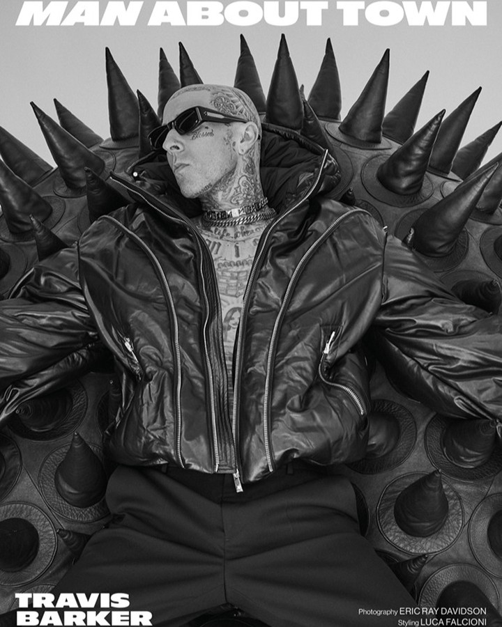Black and white studio portrait of Travis Barker in a puffer jacket, seated on a studded sphere, wearing sunglasses and chains.
