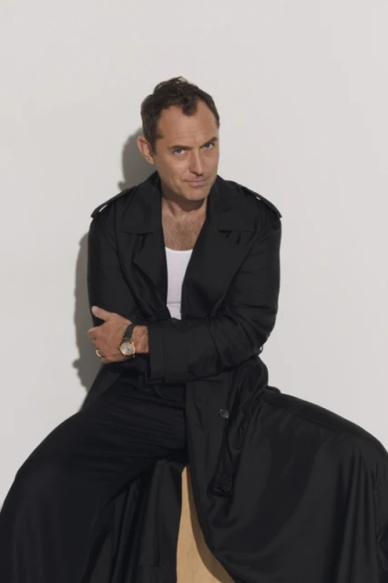 Portrait of Jude Law in a black trench coat, seated on a wooden stool in front of a white backdrop in a photo studio.