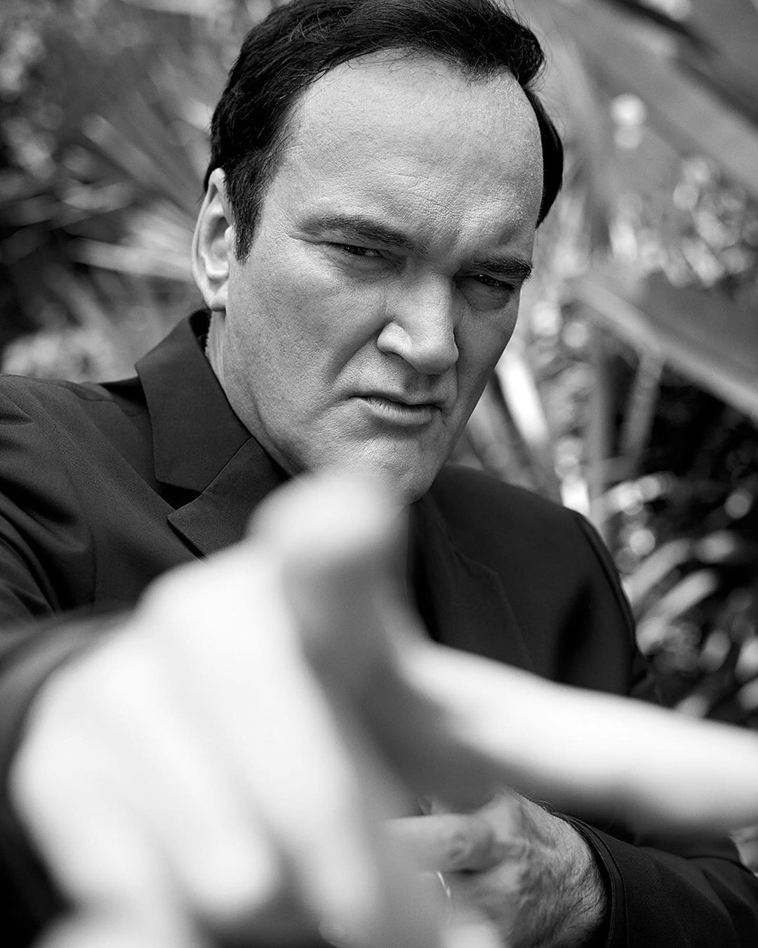 Black and white portrait of a man in a suit pointing directly at the camera, with foliage visible in the background creating depth.