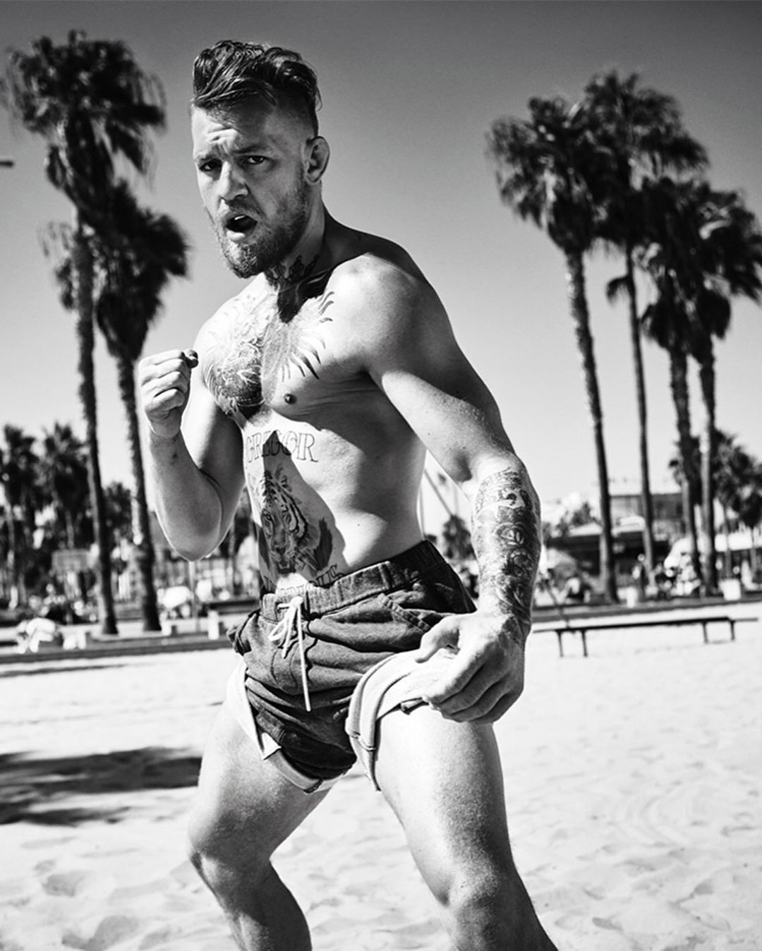Black and white portrait of bare-chested man with tattoos on a beach, possibly Conor McGregor, set against palm trees and blue sky.