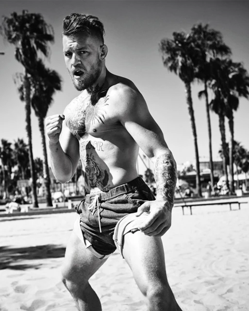 Black and white portrait of bare-chested man with tattoos on a beach, possibly Conor McGregor, set against palm trees and blue sky.