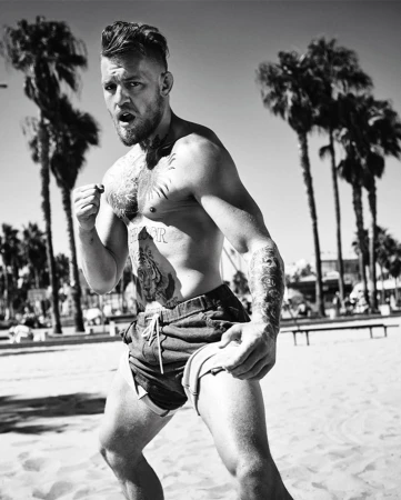 Black and white portrait of bare-chested man with tattoos on a beach, possibly Conor McGregor, set against palm trees and blue sky.