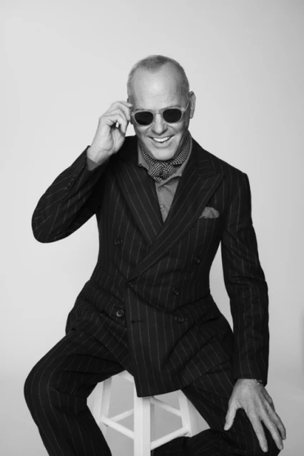 Black and white portrait of an adult male sitting on a white stool. He wears sunglasses and a pinstripe suit, smiling in the studio.