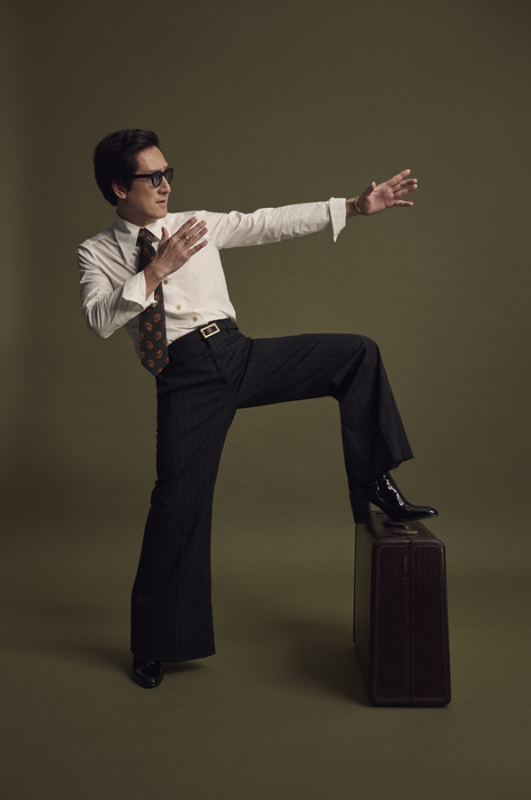 Adult male in a white dress shirt and black bell-bottom pants with glasses and vintage tie in a studio, one leg up on a suitcase.