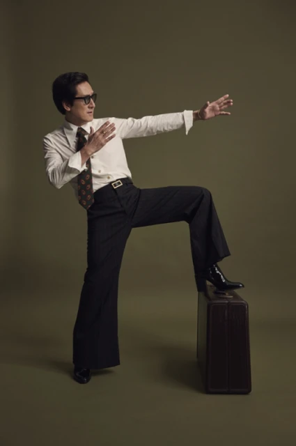 Adult male in a white dress shirt and black bell-bottom pants with glasses and vintage tie in a studio, one leg up on a suitcase.