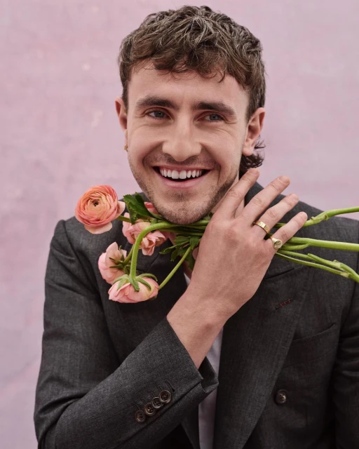 Smiling adult male in a suit, holding a bouquet of peach-colored flowers against a mottled pink background in a medium studio shot.