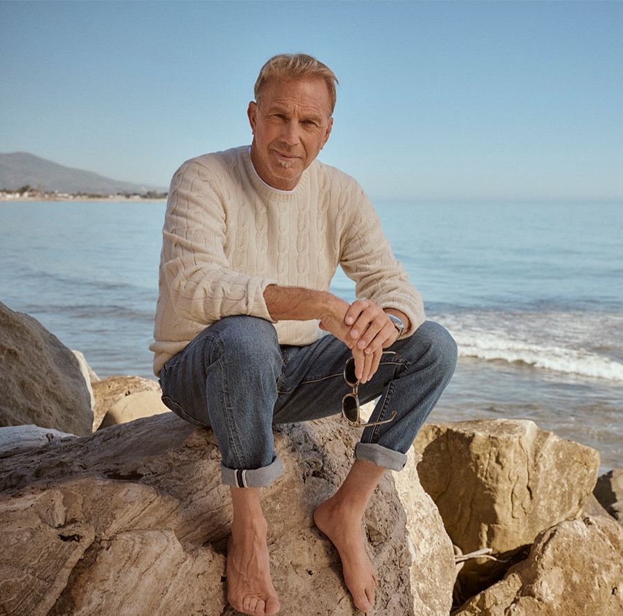 Mature man, Kevin Costner, sitting on rocks near the beach wearing a cream sweater and jeans enjoying the outdoors on a sunny day.