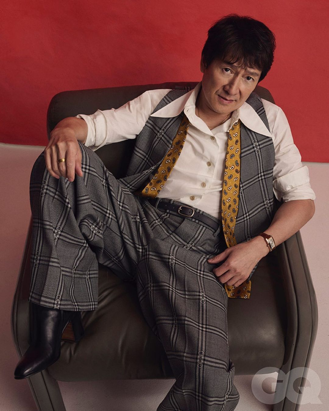 Adult man in studio reclines in chair, wearing a gray plaid suit, white button-down shirt, patterned yellow tie, and black boots against red background.