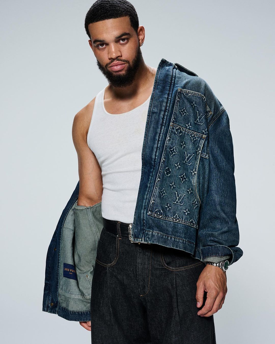 Man wearing a white tank top and dark denim pants with a monogrammed denim jacket draped over his shoulder.