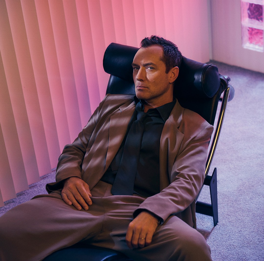Portrait of an adult man in a brown suit and black tie, sitting in a black chair indoors with pink lighting and a neutral expression.