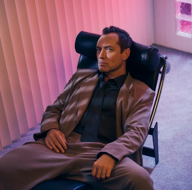 Portrait of an adult man in a brown suit and black tie, sitting in a black chair indoors with pink lighting and a neutral expression.