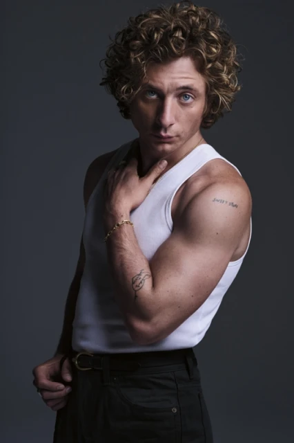 Portrait of a man with curly hair wearing a white tank top and dark pants, captured in a studio setting against a neutral background.