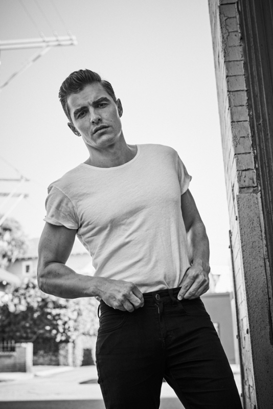 Black-and-white portrait of a muscular adult male model in a white t-shirt and black pants, shot from a low angle outdoors.