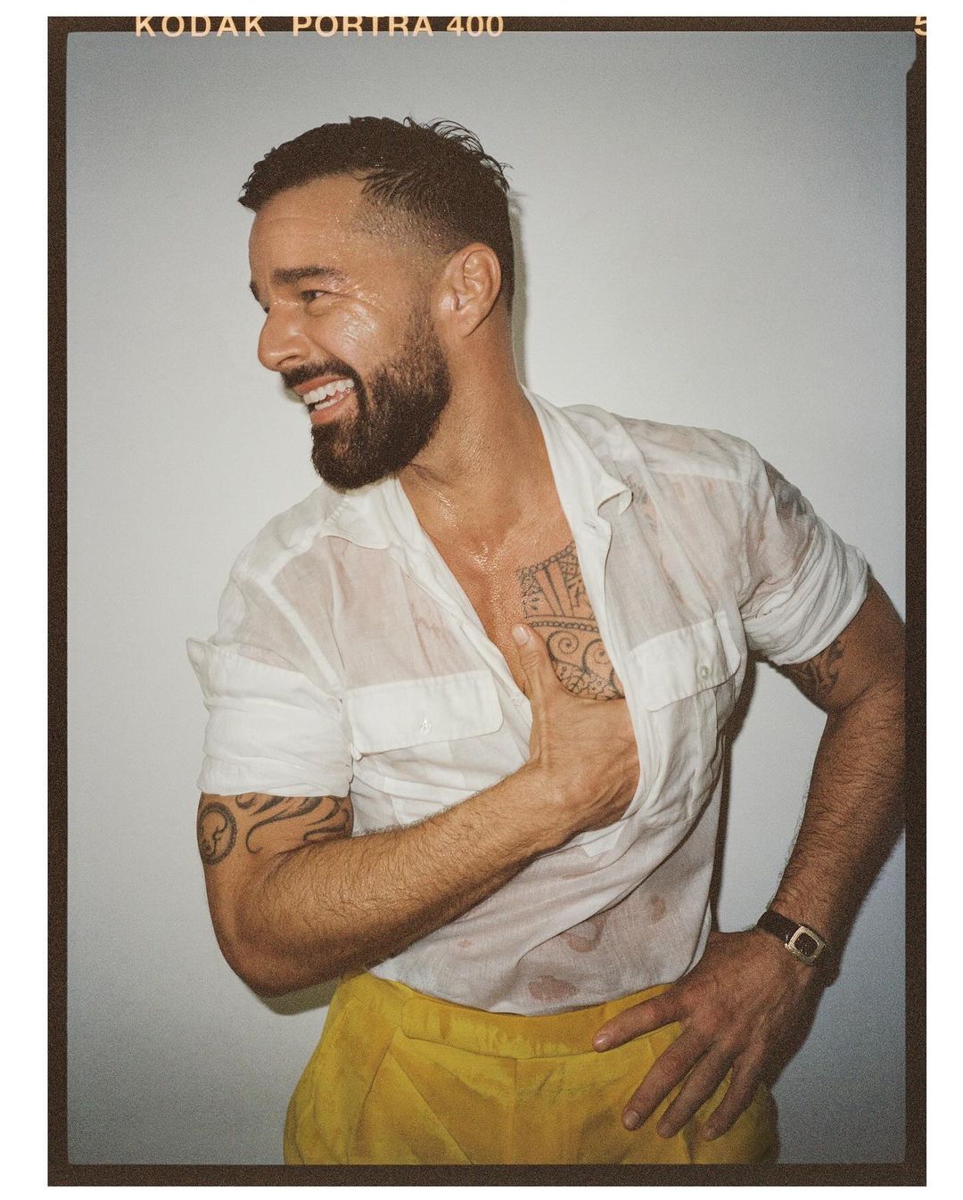 Smiling adult man in a wet white shirt and yellow pants, posing indoors with a hand on his hip and forearm tattoos visible.