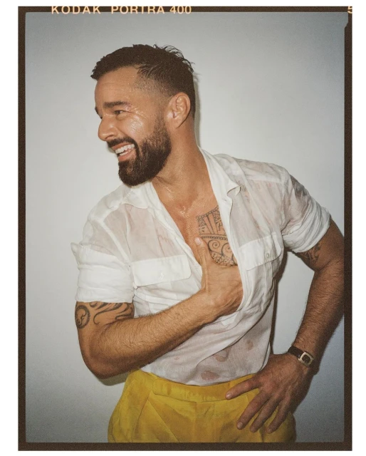 Smiling adult man in a wet white shirt and yellow pants, posing indoors with a hand on his hip and forearm tattoos visible.