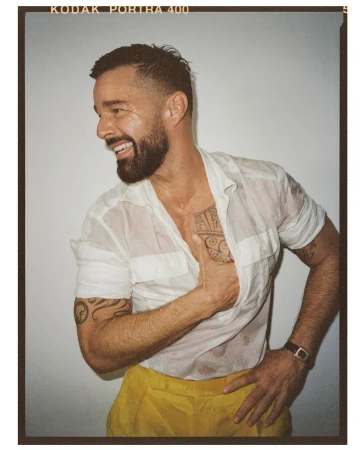 Smiling adult man in a wet white shirt and yellow pants, posing indoors with a hand on his hip and forearm tattoos visible.