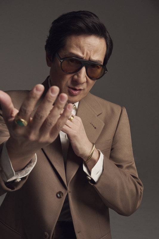 Portrait of an Asian man in a brown suit and sunglasses, holding his hand toward the camera, wearing rings and a bracelet.