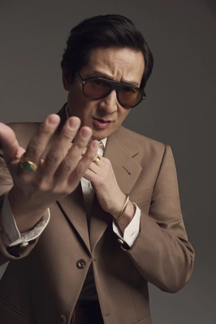 Portrait of an Asian man in a brown suit and sunglasses, holding his hand toward the camera, wearing rings and a bracelet.