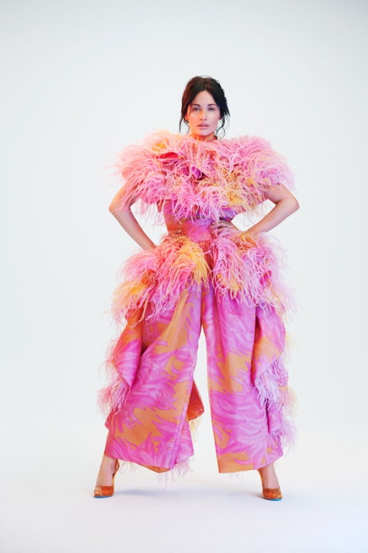 Woman wearing a voluminous pink feathered top and wide trousers standing against a white seamless background