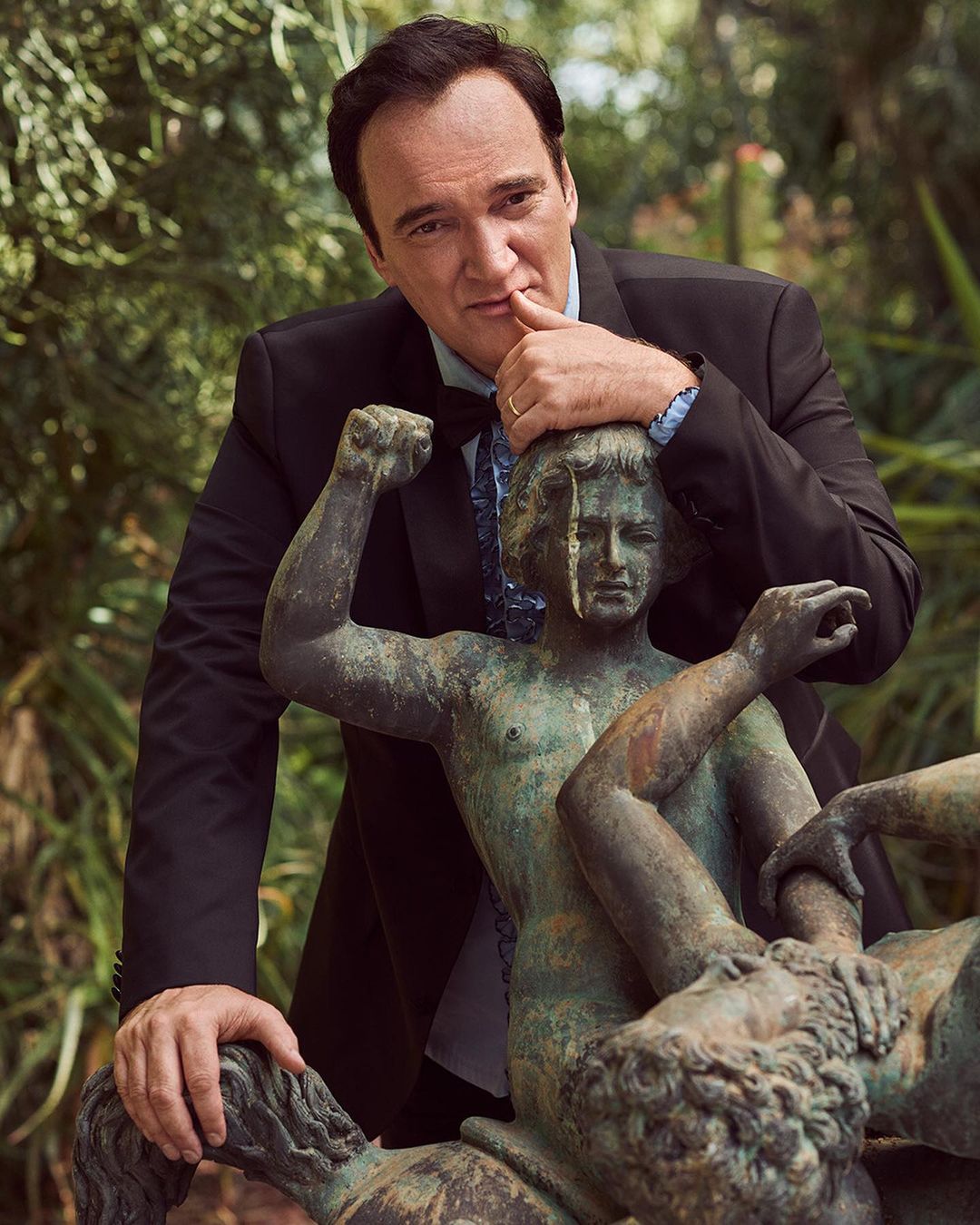 Portrait of a man in a suit posing with a weathered bronze statue of children in an outdoor setting with green foliage.