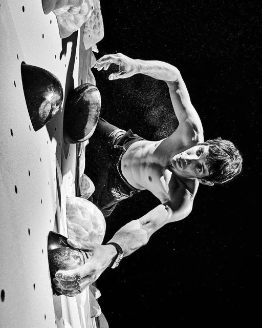 Black and white portrait of a shirtless man rock climbing on a wall, showcasing his muscular build and athletic skill against a dark background.