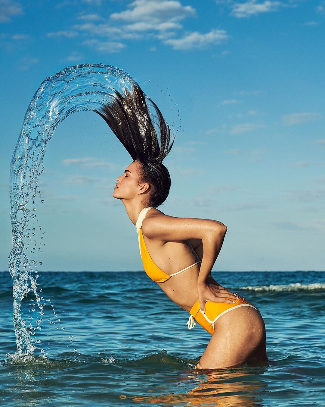 A woman stands in shallow ocean water, wearing a yellow bikini, arched back, hair flipped up creating a water halo against a blue sky.
