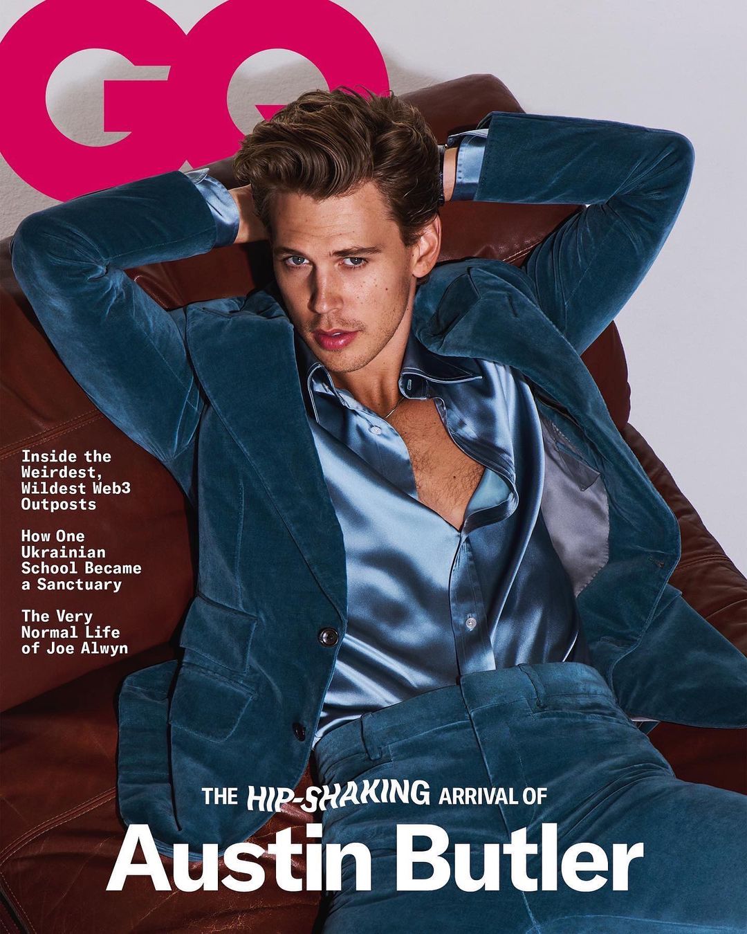 Austin Butler poses in a teal velvet suit and a silk shirt on a brown leather chair for a magazine cover, captured in a studio shot.