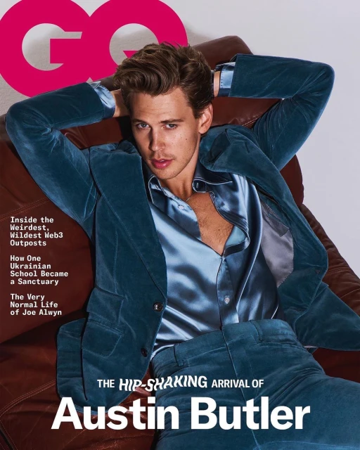 Austin Butler poses in a teal velvet suit and a silk shirt on a brown leather chair for a magazine cover, captured in a studio shot.