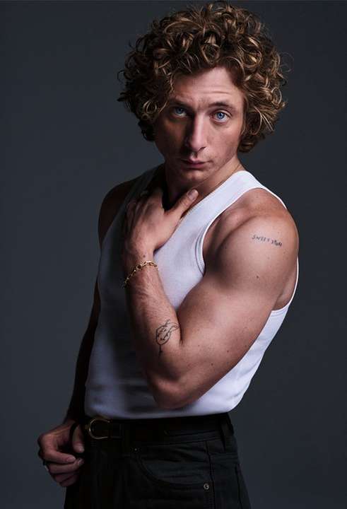 Indoor studio portrait of a blonde curly-haired adult male wearing a white tank top and dark pants, posing with his hand near his chest.