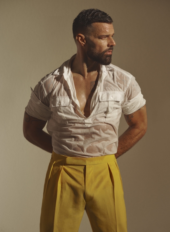 Attractive man in wet white shirt and yellow pants poses in a studio with arms behind his back and looking right, against beige background.