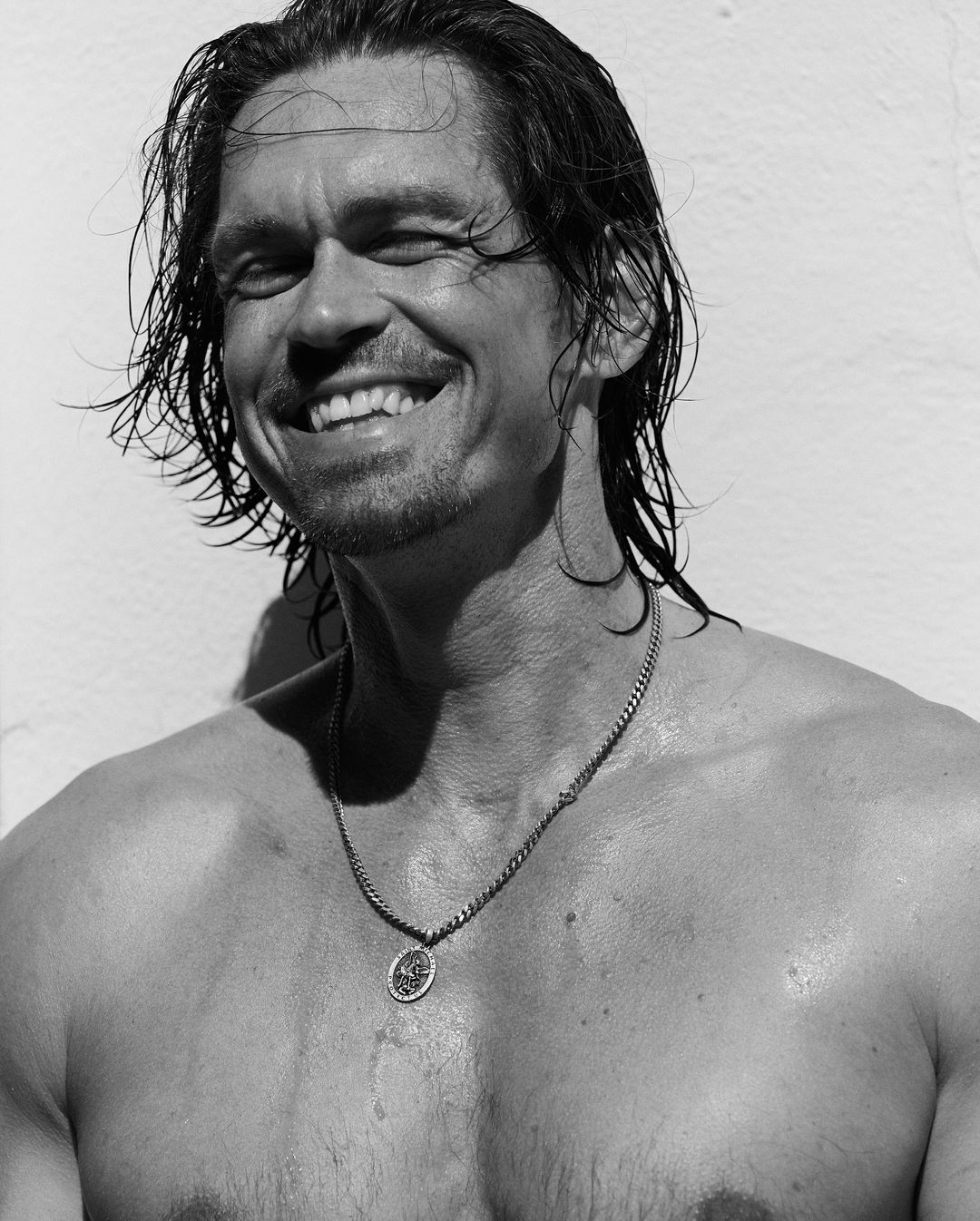Close up monochrome portrait of a smiling man with wet hair wearing a silver chain necklace against a light wall