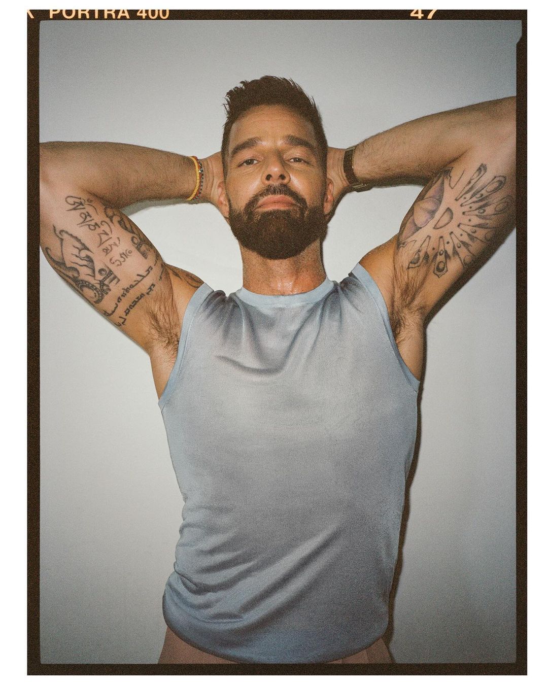 Portrait of adult male with arm tattoos posing in front of a white wall, wearing a tank top and bracelet with a neutral expression.