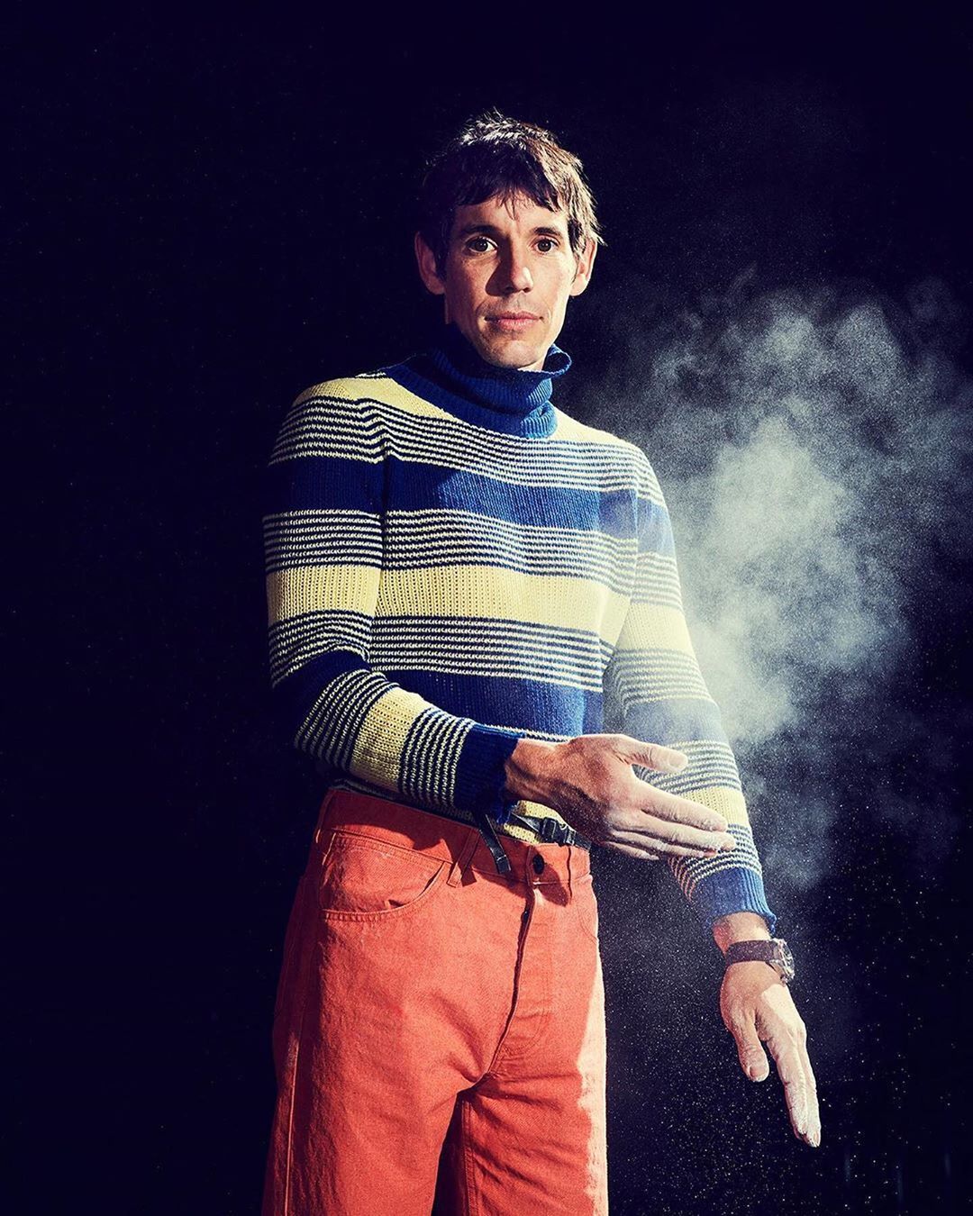 Full studio shot of an adult male wearing a striped turtleneck and orange pants, with white chalk on hands against a black backdrop.